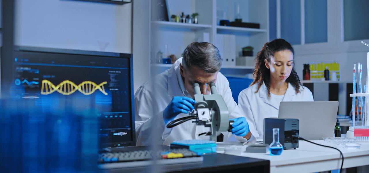 One researcher working with a microscope while the other person documenting on a laptop.