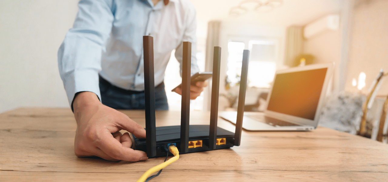 Person adjusting a Wi‑Fi router with while holding a smartphone, with a laptop on a wooden desk in the background.