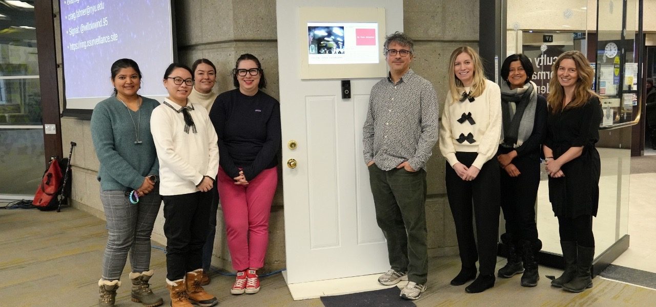A group of people stands beside a white door installation with a small screen and camera.